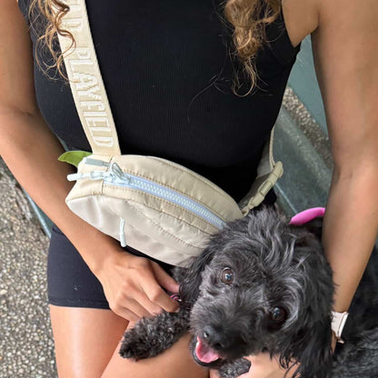 A person sitting on a bench holding a small black dog, wearing a beige crossbody bag with the brand name 'PLAYFIELD' printed on the strap.