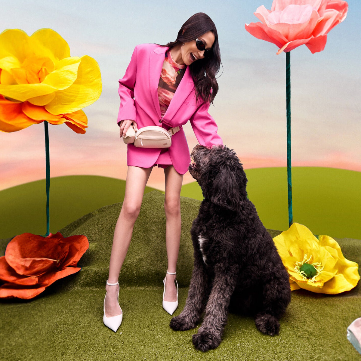 A woman in a bright pink blazer and matching mini skirt stands on artificial grass, wearing white pointed heels and holding a beige crossbody bag. She is interacting with a black poodle.